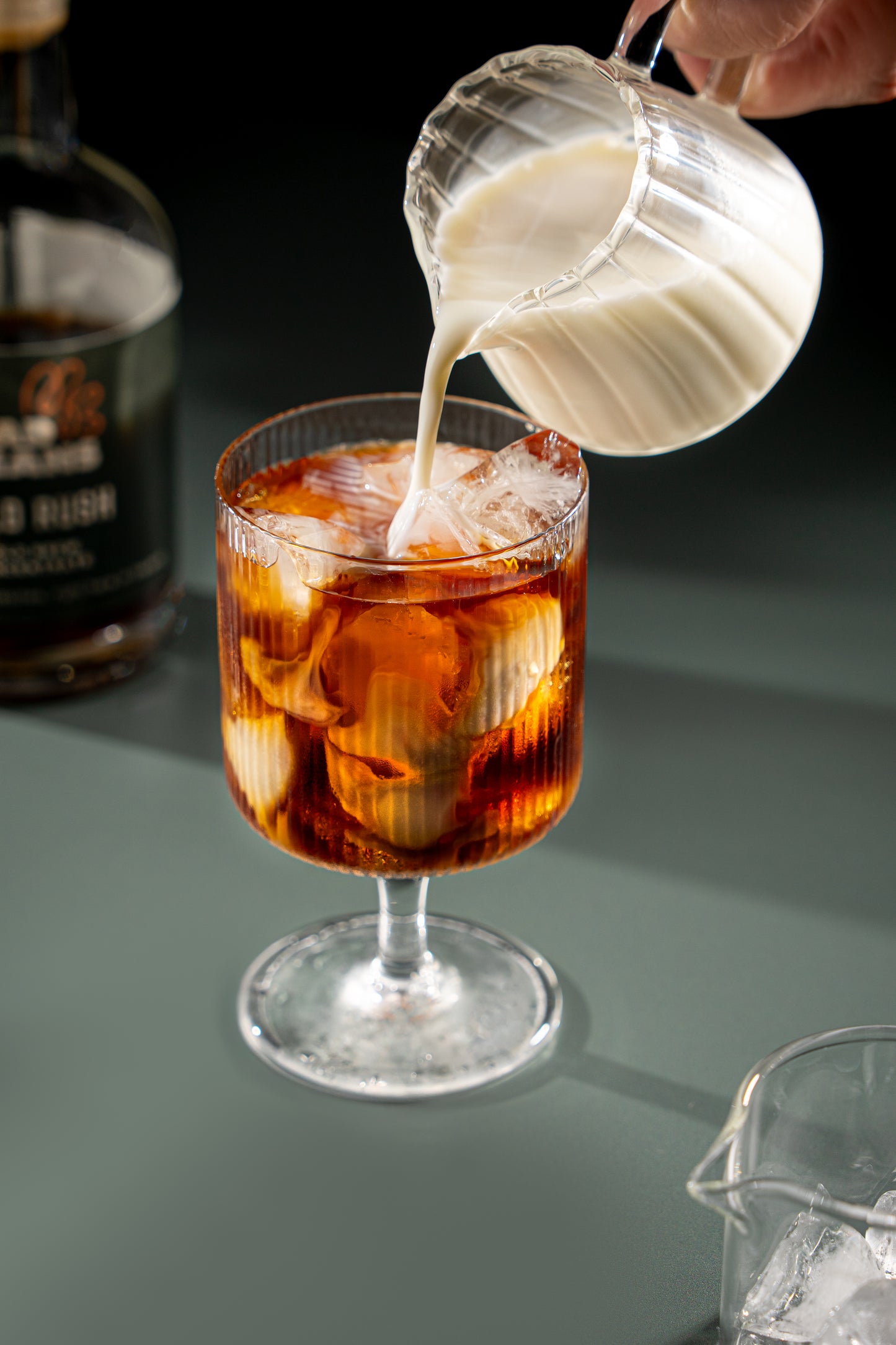 Pouring cream into a glass of iced coffee with a bottle of Mad Beans Cold Rush in the background.
